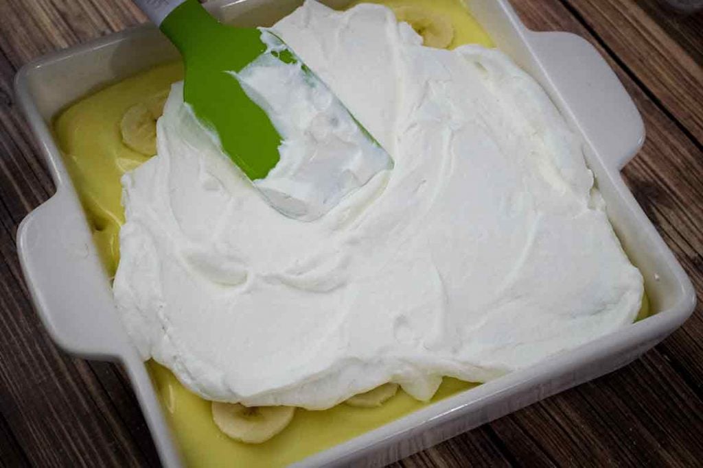 A green rubber spatula spreading whipped cream on top of the banana pudding layer.