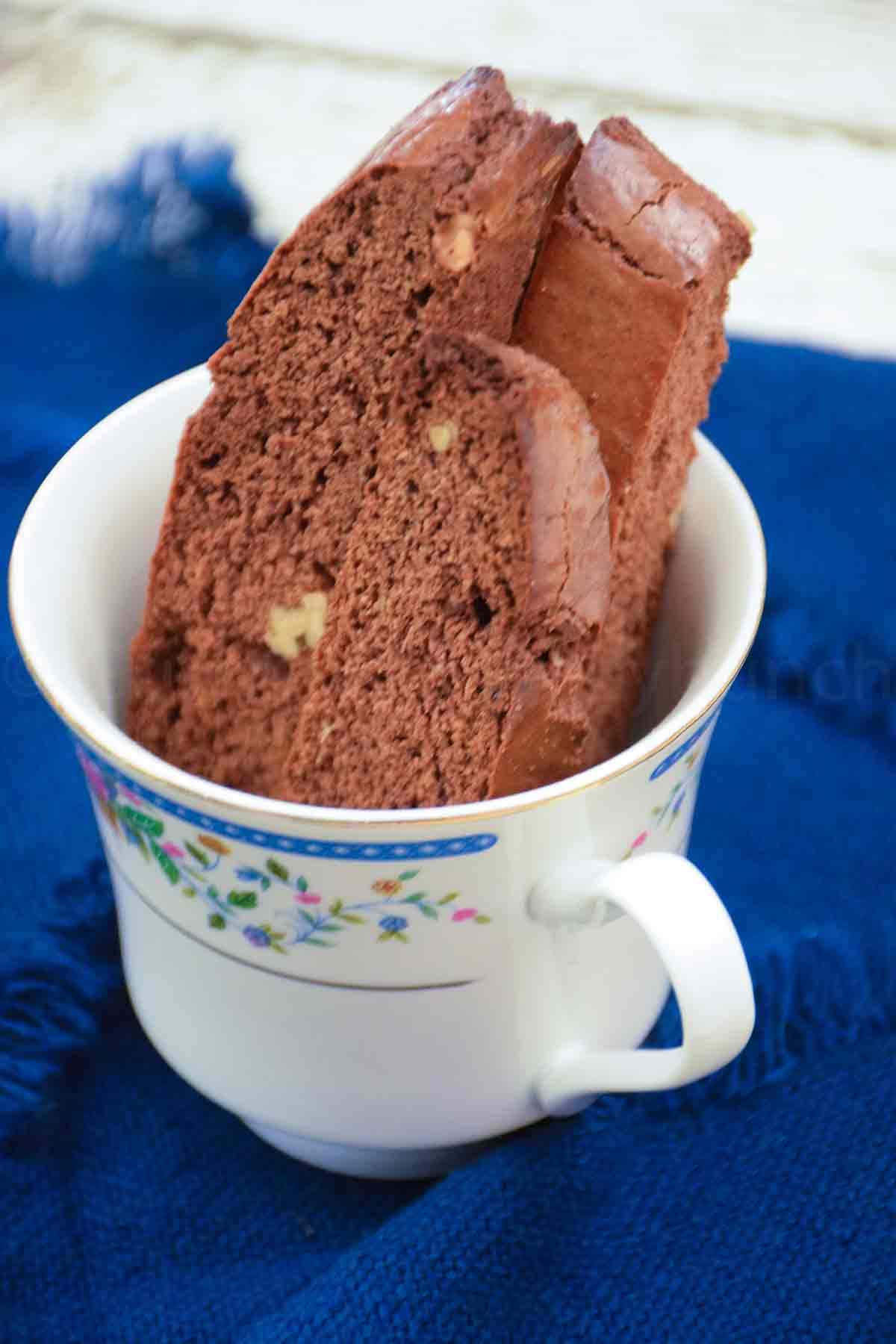 Three chocolate biscotti standing up in a china cup with a blue napkin next to the cup.