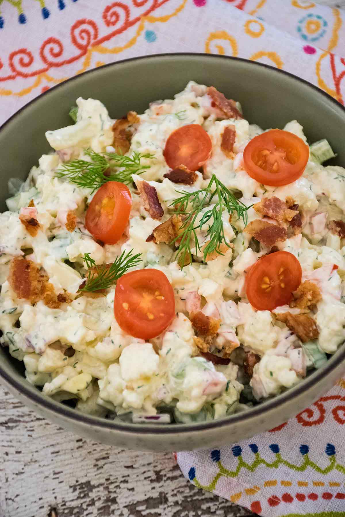 A close-up of keto faux potato salad made with cauliflower, topped with halved cherry tomatoes, crispy bacon pieces, and fresh dill sprigs, served in a speckled bowl on a green patterned napkin.