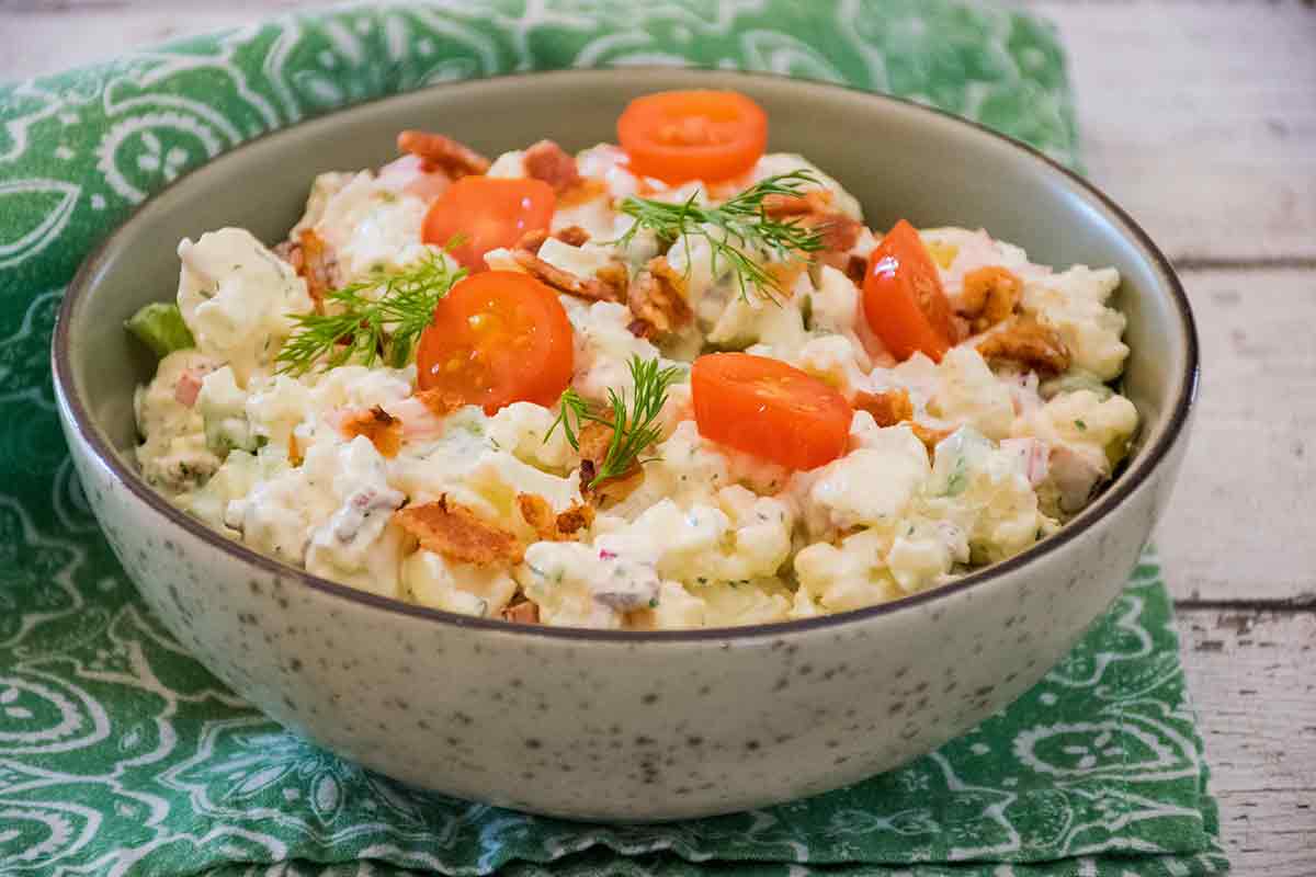 A bowl of creamy faux potato salad made with cauliflower, garnished with cherry tomato slices, crispy bacon bits, and fresh dill sprigs, set on a red and brown patterned cloth.