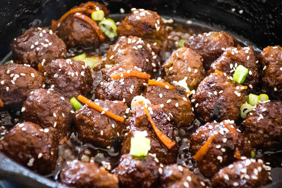 Mongolian meatballs simmering in a glossy, dark sauce inside a skillet, garnished with sliced green onions, carrot strips, and sesame seeds.
