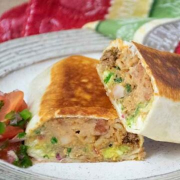 A grilled stuffed burrito cut in half on a plate, showing a filling of beef, beans, guacamole, and melted cheese, served with a side of fresh salsa.