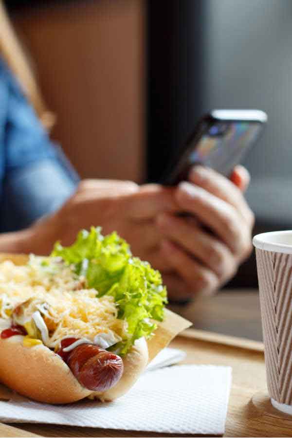 a photo of a hotdog and cup of coffee with person in the background holding a phone (distracted eating)