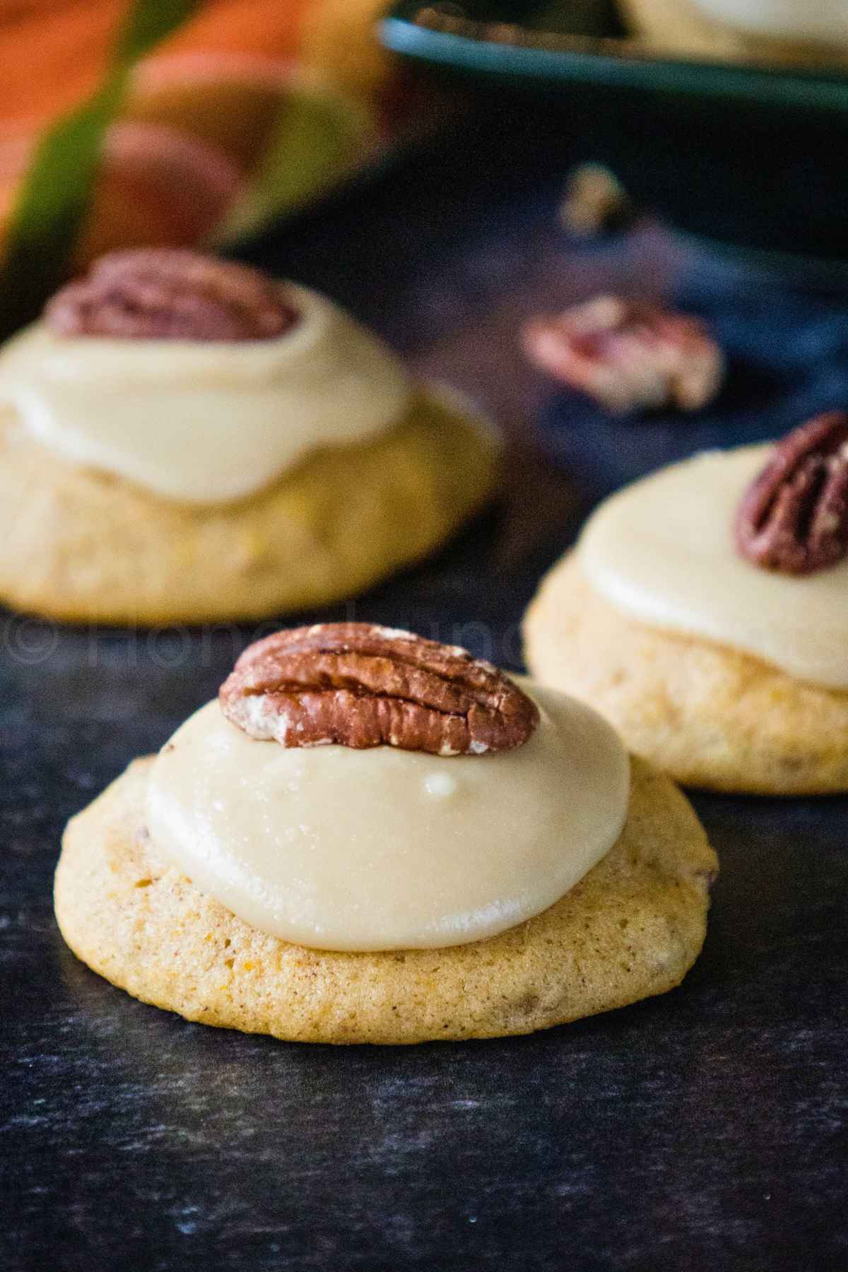 Soft Butternut Squash Cookies topped with creamy brown sugar frosting and a pecan half, arranged on a dark surface.