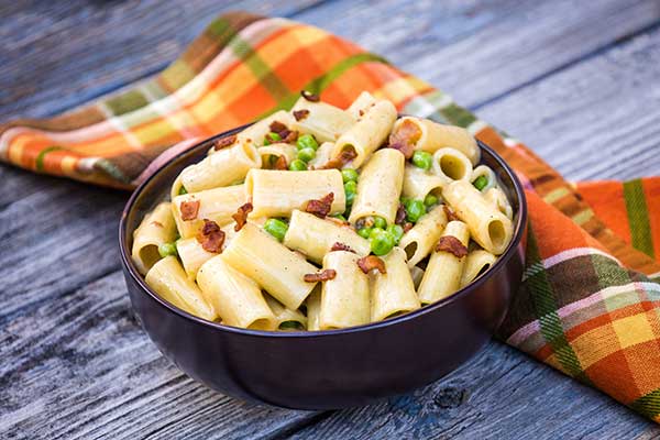 A bowl of one pot pasta sprinkled with crumbled cooked bacon and green peas.