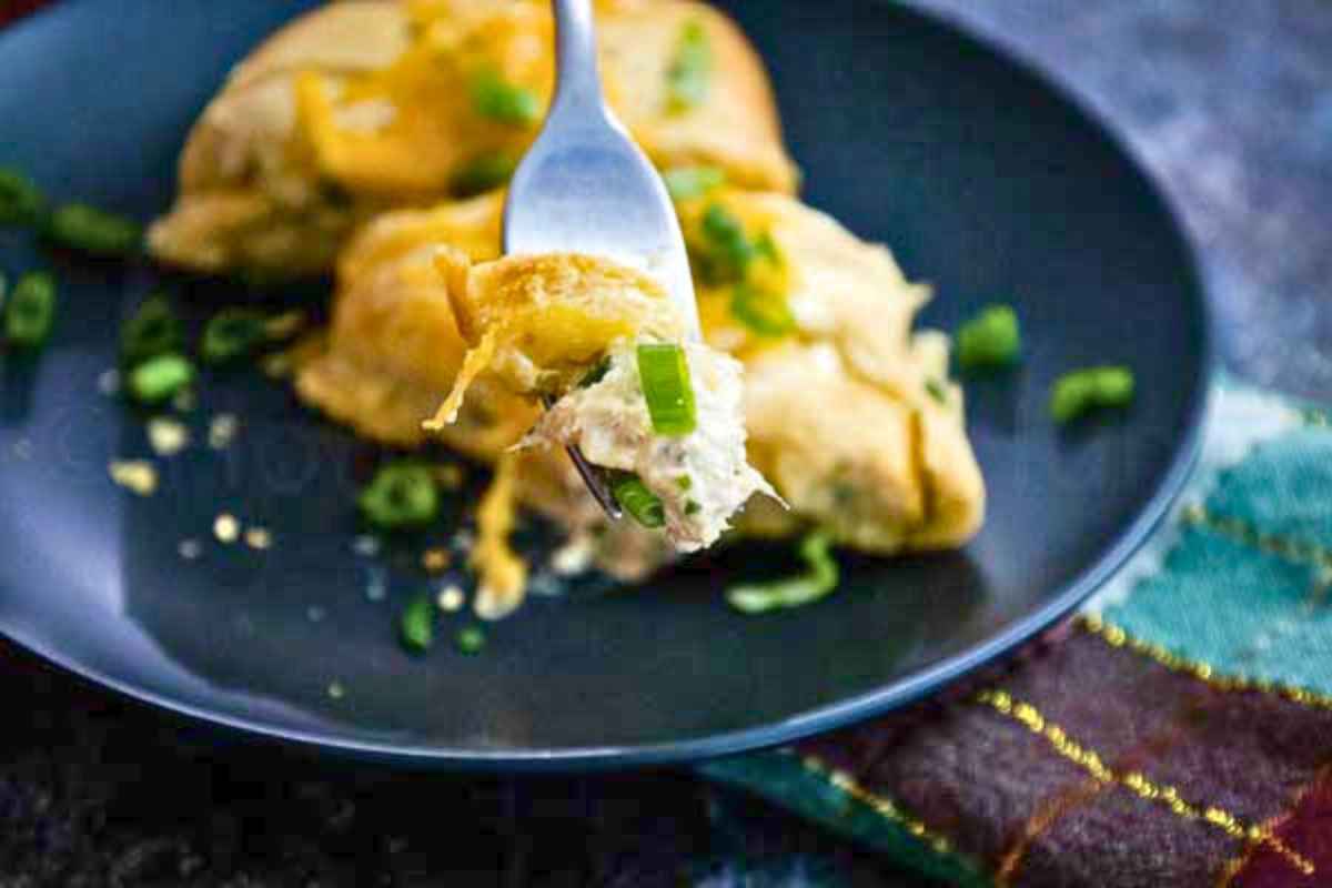 A fork lifts a bite of cheesy tuna crescent braid topped with melted cheddar and fresh green onions from a black plate.