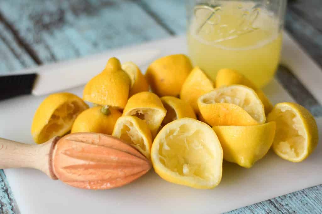 Squeezed lemons on a white cutting board with a knife and pint jar of juice in the background and a wooden citrus reamer in the front.