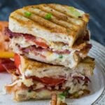 Two halves of a crispy grilled artisan bread sandwich stacked on a white plate, showing melted cheese, thick tomato slices, and bacon.