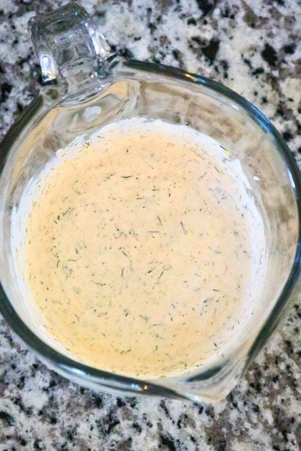 Glass measuring cup filled with creamy dill sauce, speckled with herbs, sitting on a gray and black speckled countertop.