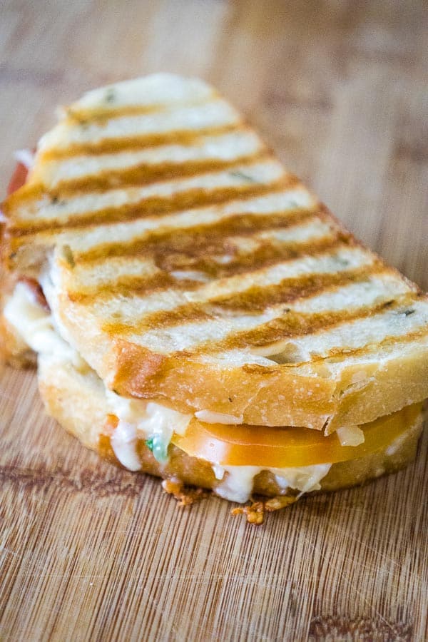 A close-up of a grilled artisan bread sandwich with visible grill marks, filled with melted white cheese, a thick slice of Hierloom tomato, bacon, and fresh scallions.