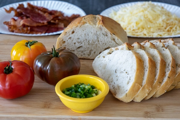 A wooden cutting board with a fresh loaf of artisan bread, thick slices of raw heirloom tomatoes, a bowl of sliced green onions and a plate of cooked bacon.