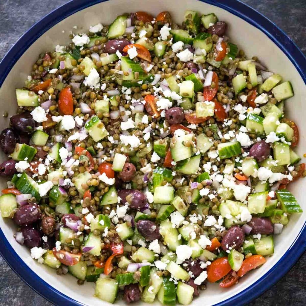 Overhead view of a fully assembled lentil salad made with brown lentils, black kalamata olives, red grape tomatoes, diced cucumbers and red onions tossed with feta cheese in a cream ceramic serving dish with a blue rim.