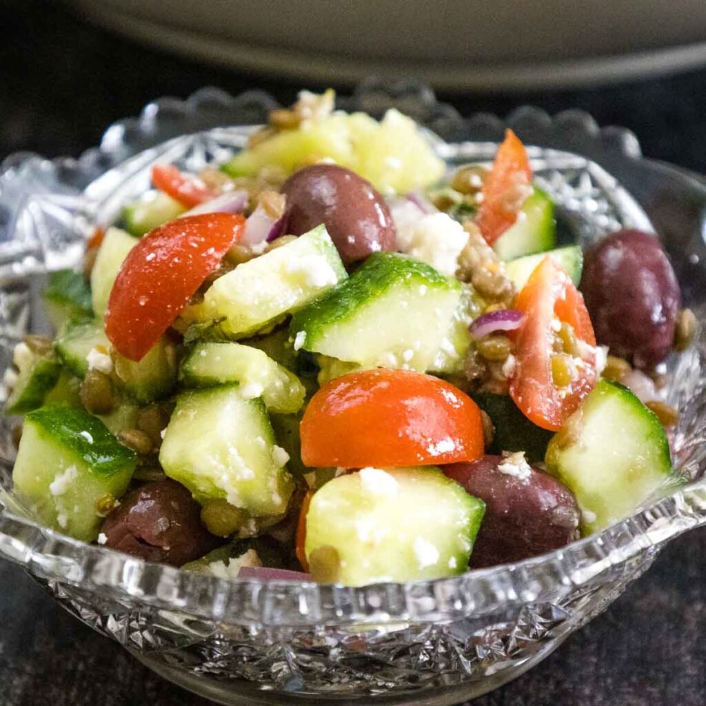 Crystal glass bowl with a single serving of lentil salad made with diced cucumbers, grape tomatoes, black olives, feta cheese in a lemon dressing.