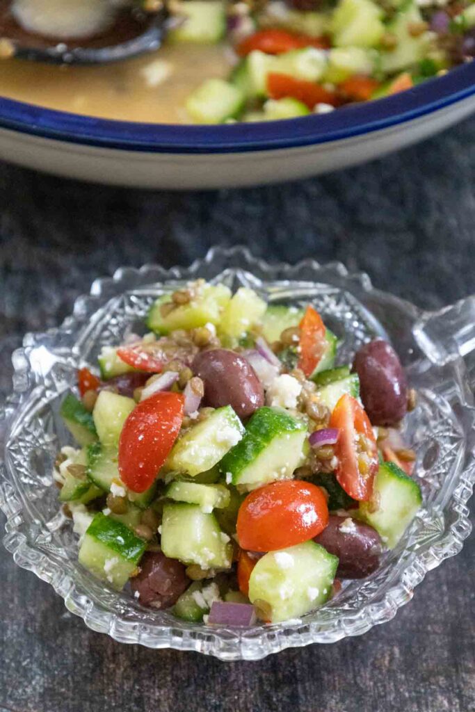 A single serving of salad made with cucumbers, grape tomatoes, red onion, feta cheese, black olives, and cooked lentils in a glass serving dish.