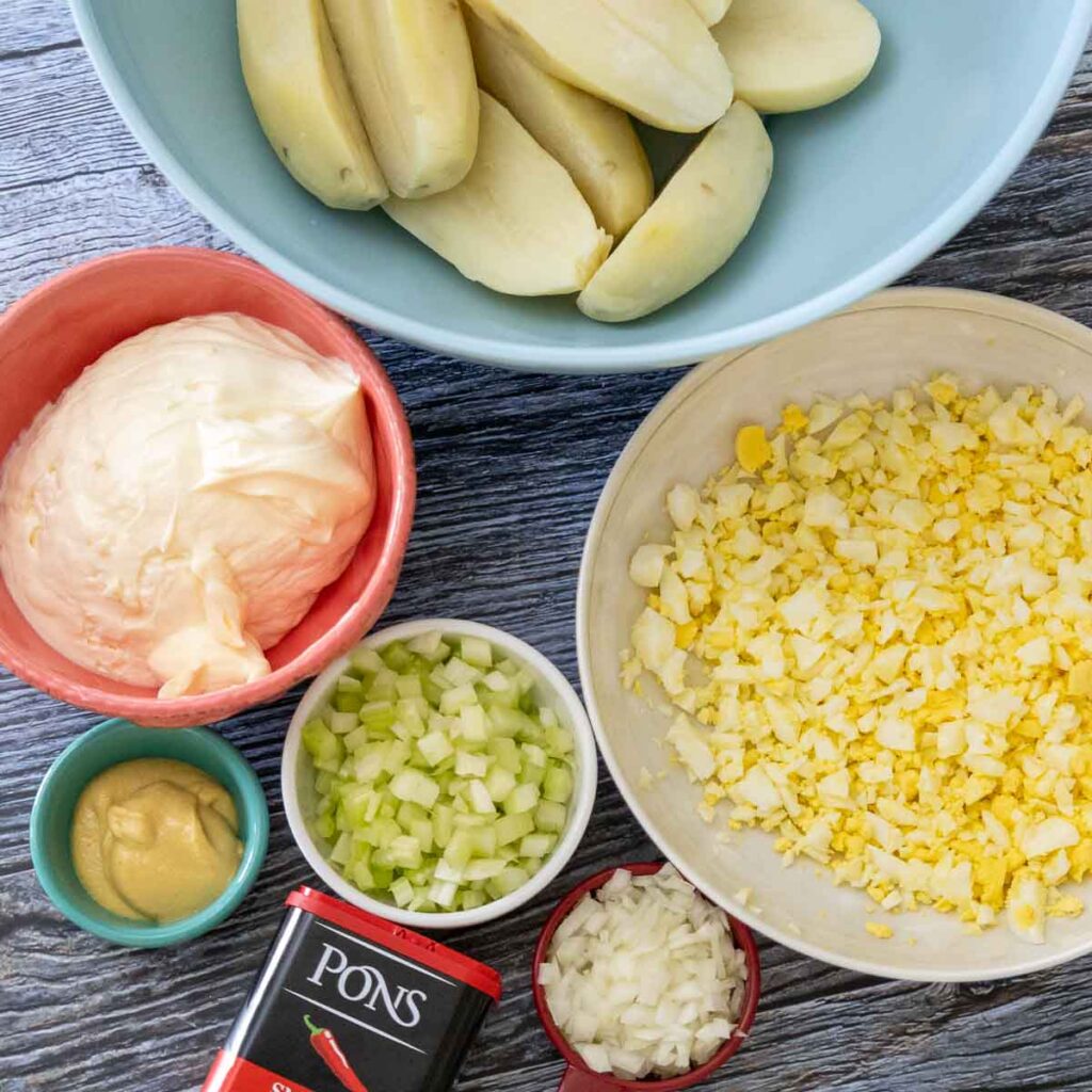 Potato Salad Ingredients clockwise, cooked potatoes in a blue bowl, chopped hard boiled eggs in a cream colored bowl, a red measuring cup with diced onions, a can of smoked paprika, a white dish with diced celery, a turquoise dish with yellow mustard, and a pink bowl with mayonnaise.