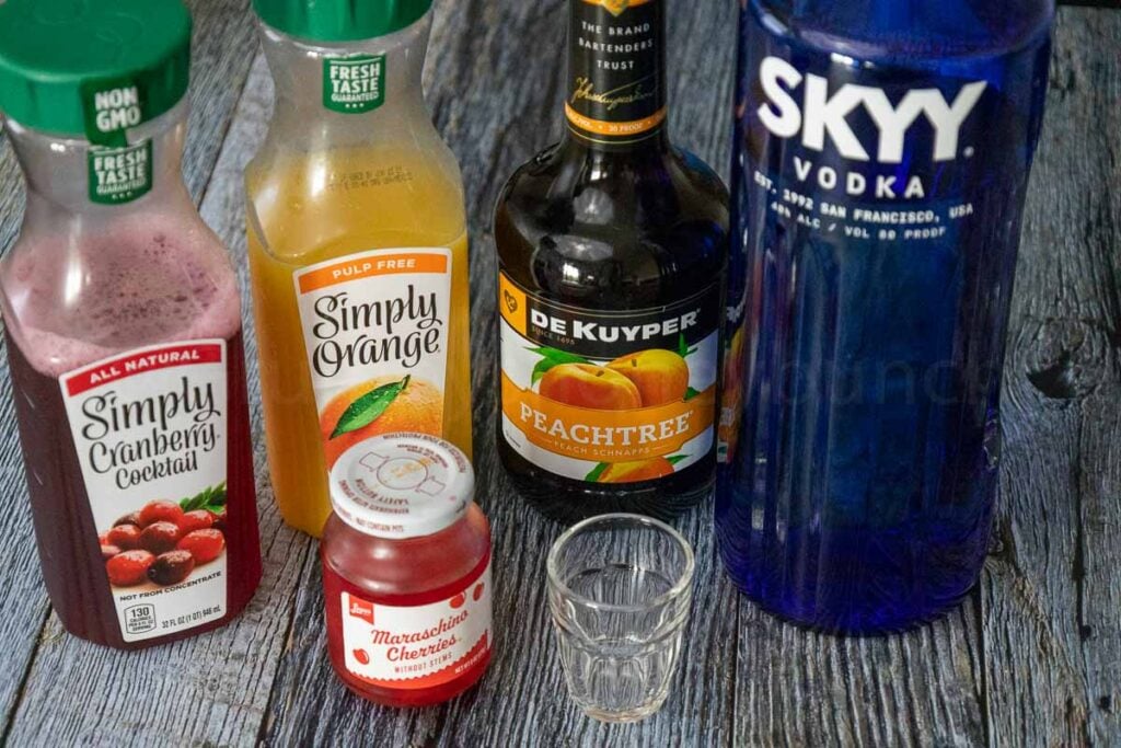 A line up of the ingredients from back to front, left to right: Cranberry Juice, Orange Juice, Peach Schnapps and Vodka. In front, Maraschino Cherries and a shot glass.