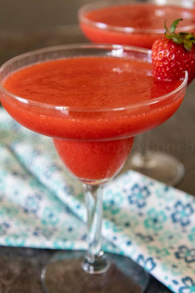 A frozen strawberry daiquiri garnished with a whole fresh strawberry with a white and blue flowered napkin between the glass blurred.