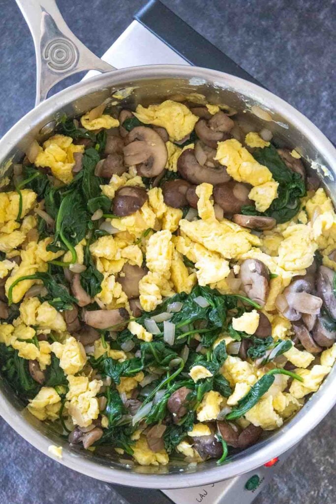 Stainless Steel Skillet with scrambled eggs, mushrooms, onion pieces, and spinach combined together.