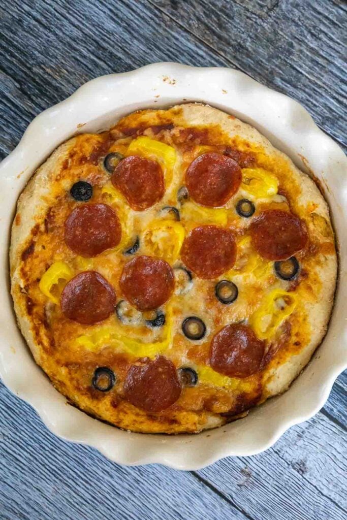 Overhead view of one pan pepperoni pizza in a white ceramic dish with pepperoni, banana peppers, and black olive toppin a golden brown crust.