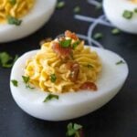 Close-up of a deviled egg topped with piped filling, bacon bits, and chopped parsley on a dark serving platter.