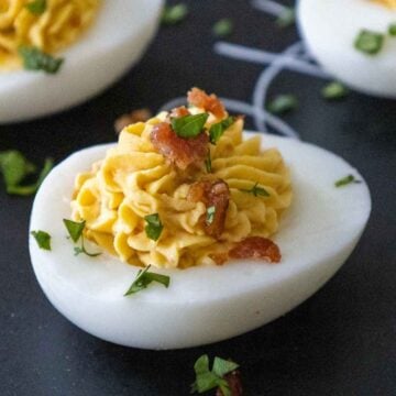 Close-up of a deviled egg topped with piped filling, bacon bits, and chopped parsley on a dark serving platter.