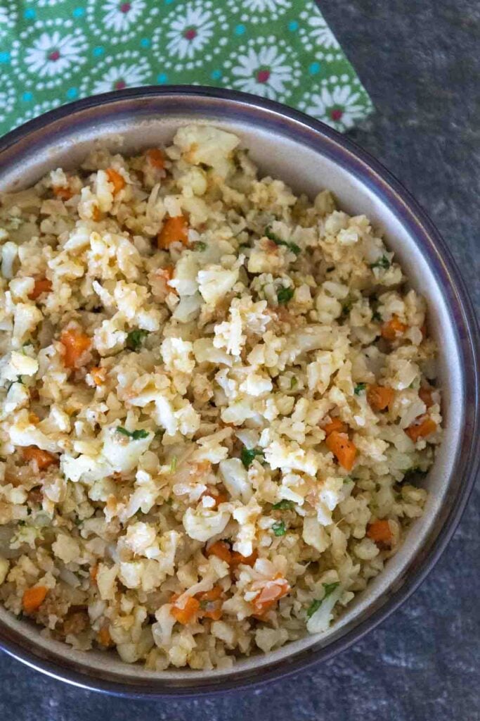 Overhead image of cauliflower rice in a beige bowl with dark brown rim and flecks of orange carrot and green cilantro throughout the rice.