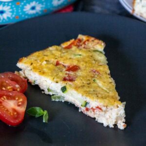 A wedge of keto zucchini pie on a black plate with a sliced cherry tomato on the side garnisheed with fresh thyme.