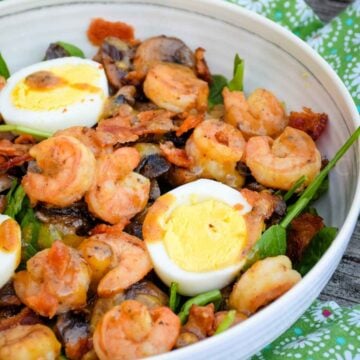 A top-down view of a healthy Shrimp Spinach Salad served in a blue bowl, garnished with sliced hard-boiled eggs and a warm bacon dressing.