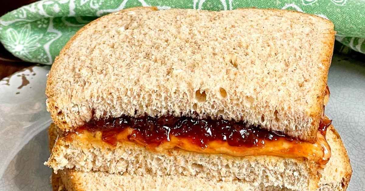 A classic peanut butter and. muscadine jam sandwich made with soft whole wheat bread, cut in half and stacked on a plate. In the background, a jar of muscadine jam with a spoon inside sits next to a patterned green cloth.