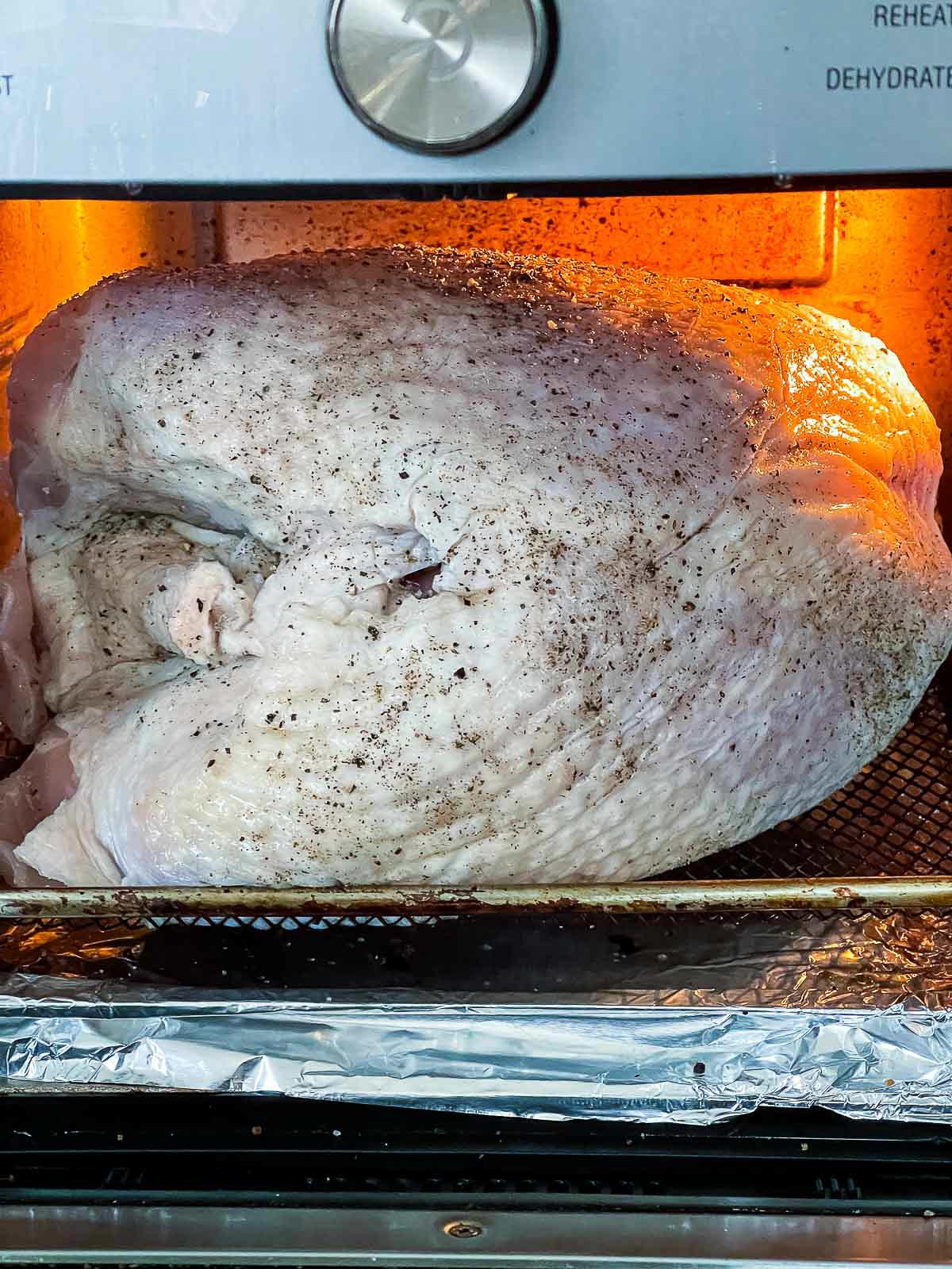 Raw seasoned turkey breast inside an air fryer oven beginning to cook under warm interior lighting with visible black pepper seasoning on the skin.