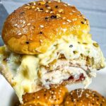 A hot turkey cranberry slider being lifted on a spatula, with melted cheese, turkey, stuffing, and cranberry sauce inside a golden, seeded bun.