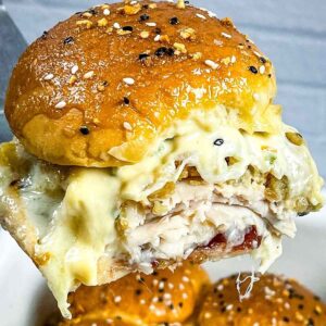 A hot turkey cranberry slider being lifted on a spatula, with melted cheese, turkey, stuffing, and cranberry sauce inside a golden, seeded bun.