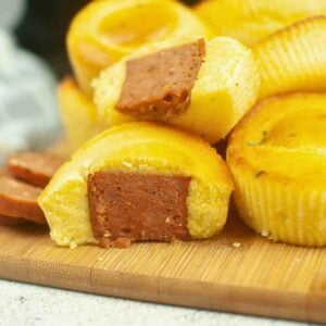 Upclose image with golden brown cornbread muffins, some with visible sausage centers resembling mini sausage corn dog bites, with sliced sausage scattered around.