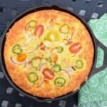 Overhead image of jalapeno cornbread baked in a cast iron skillet with red onion, jalapeno, and halved cherry tomatoes baked on top.