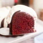 A square image with a slice of red velvet bundt cake topped with cream cheese icing on a cake serve.