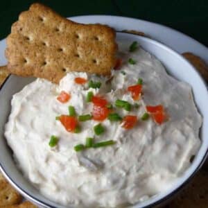 A bowl of creamy clam dip with chive and pimento topping and a whole wheat cracker in the top of the dip.