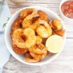 Overhead image of crispy shrimp in a white bowl with a lemon wedge on the side and a small white bowl of cocktail sauce.