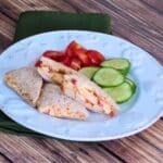 Pimento Cheese Tea Sandwiches cut into triangle shaped quarters on a plate with sliced cucumbers and tomatoes.