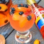 A glass of peach sangria in a clear footed glass with fresh peach and blueberries in the drink.