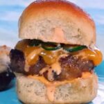 Upclose image of a sausage burger on a slider roll topped with cheddar cheese, jalapeno slices and sriracha aioli.