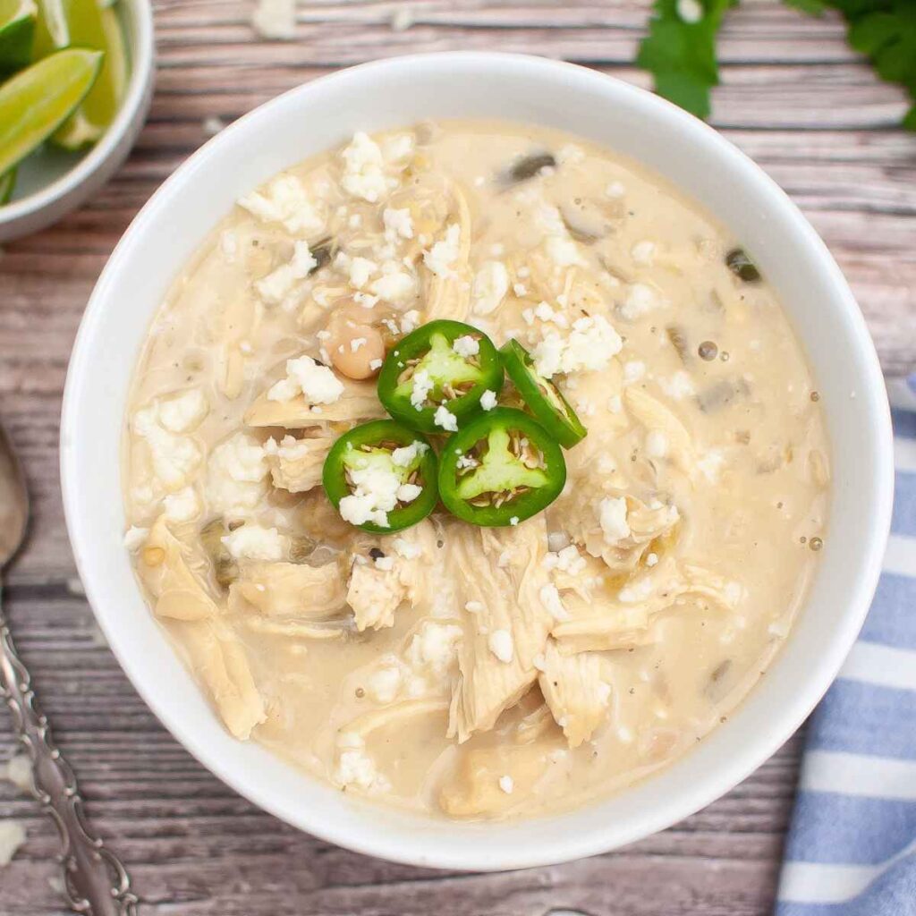 Overhead image of a white bowl full of green chicken chili topped with jalapeno slices and crumbled cheese.