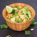 A wooden bowl filled with chicken noodle salad, topped with chunks of chicken, cucumber slices, shredded cabbage, carrots, and fresh cilantro. A lime wedge sits on top, with extra cilantro leaves scattered on the dark surface around the bowl.
