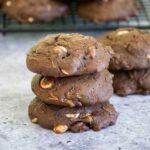 A stack of three thick, soft chocolate cookies with white chocolate chips and visible bits of zucchini, set on a parchment-lined surface. More cookies are cooling on a wire rack in the background.
