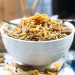 A close up side view of white textured bowl filled with creamy air fryer green bean casserole, topped with crispy fried onions. Scattered grean beans and onion pieces are visible on the white surface below the bowl, with a soft plaid napkin partially in frame.