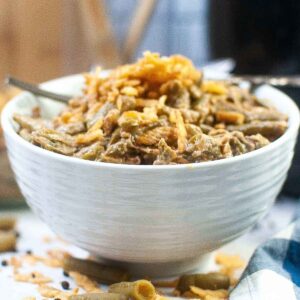 A close up side view of white textured bowl filled with creamy air fryer green bean casserole, topped with crispy fried onions. Scattered grean beans and onion pieces are visible on the white surface below the bowl, with a soft plaid napkin partially in frame.