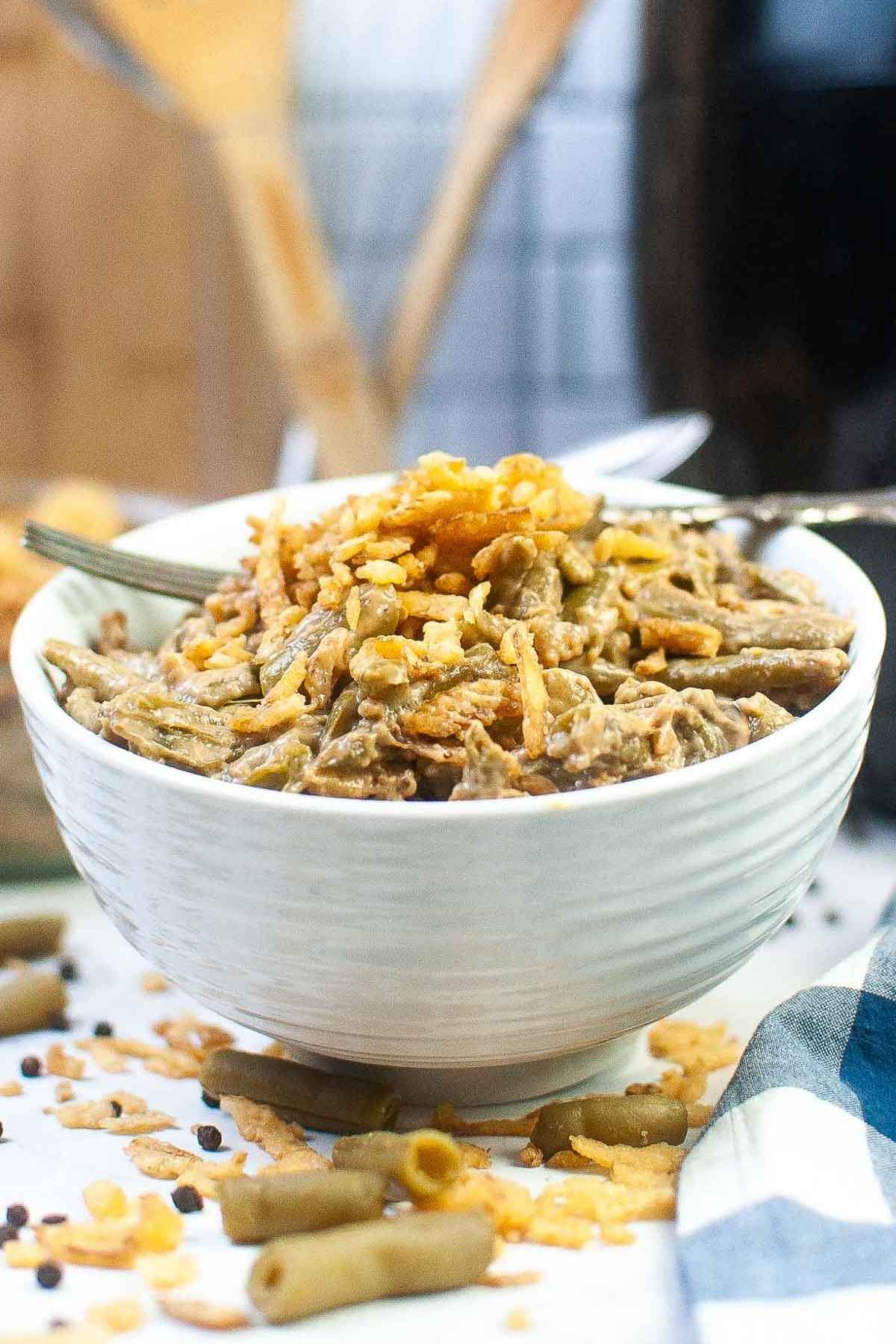 A close up side view of white textured bowl filled with creamy air fryer green bean casserole, topped with crispy fried onions. Scattered grean beans and onion pieces are visible on the white surface below the bowl, with a soft plaid napkin partially in frame.