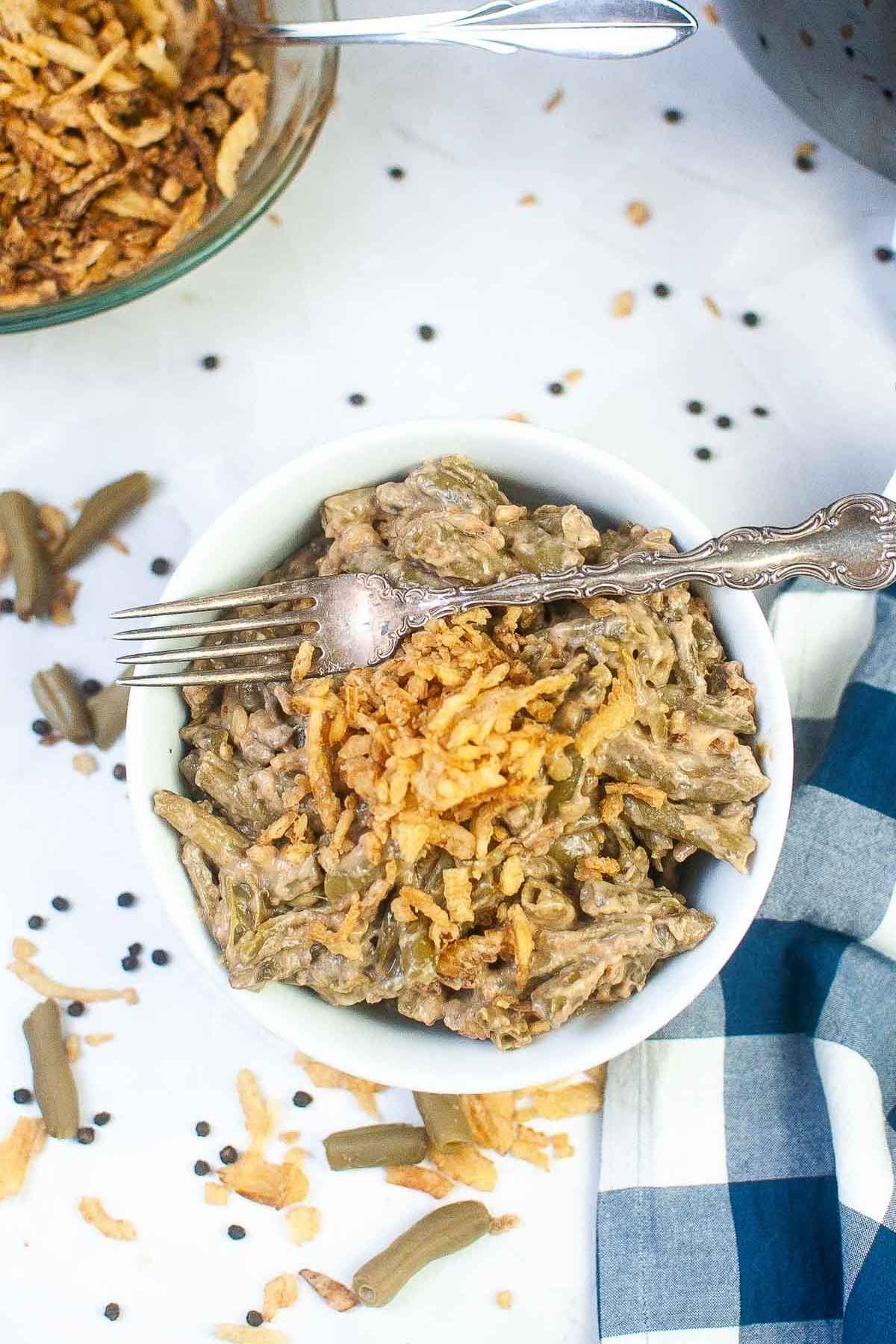 Overhead view of a bowl of green brean casserole topped with crispy fried onions and a vintage fork, surrounded by scattered black peppercorns, green beans, and onion pieces on a white surface, with a glass bowl of extra fried onions and a blue and white checkered napkin nearby.