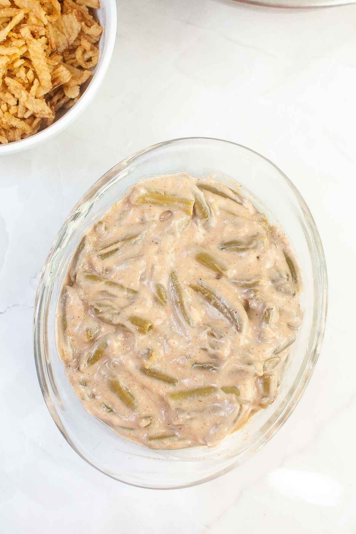 A glass bowl filled with an uncooked green bean casserole mixture made with green beans and creamy sauce, placed on a white countertop next to a bowl of crispy fried onions.