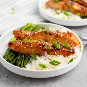 Bowl of teriyaki glazed salmon fillets served over white rice with roasted asparagus and garnished with sesame seeds and sliced green onions.