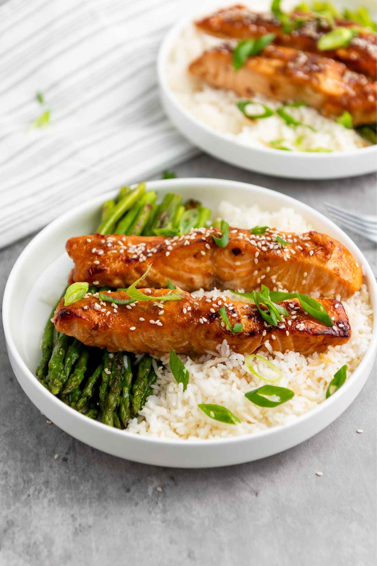 Two pieces of Tariyaki salmon served in a white bowl with rice and asparagus, garnished with sesame seeds and scallions, placed on a light gray surface.
